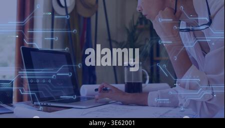 Leaning mature woman in white shirt examining laptop, plans, HUD overlay at home office, copy space Stock Photo