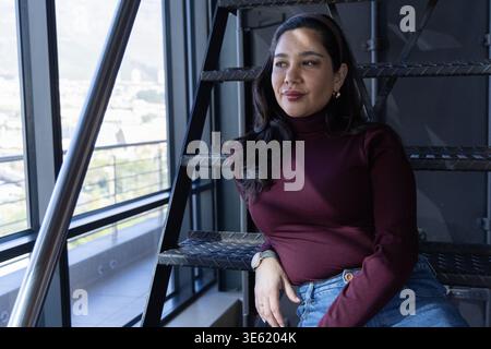Woman in her 30s sitting on diamond-plate metal stairs by large glass windows, wearing wristwatch Stock Photo