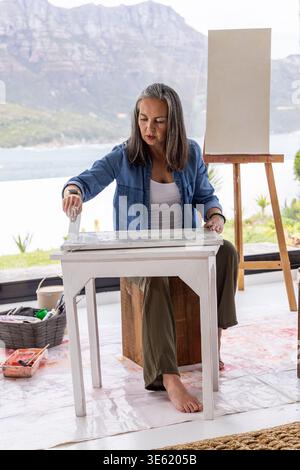 Senior female painter sitting on wooden stool priming small white table in studio with blank canvas Stock Photo