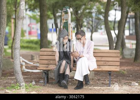 In June 2025, a Korean man and woman in their late fifties sit on a bench by the sea, chatting and smiling as they enjoy the breeze, ocean view, and c Stock Photo