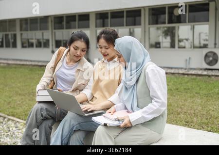 A group of diverse students working together using a laptop and study materials while sitting outdoors at a university campus Stock Photo
