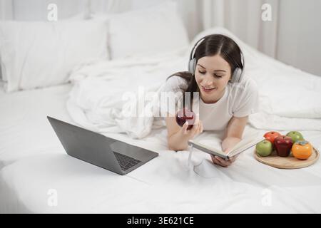 A woman relaxes in a comfortable bedroom setting, enjoying music and fresh fruits while using a laptop and reading a book Stock Photo