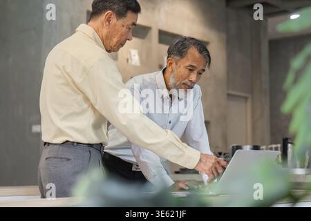 Two experienced men share ideas while reviewing data on a laptop, emphasizing collaboration and strategic planning in a modern business environment Stock Photo