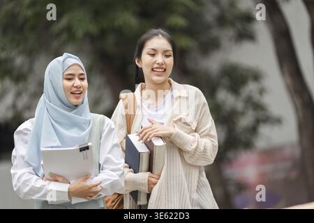 Two diverse college students, one in a hijab, enjoying their time outdoors on campus while carrying books and sharing a moment of friendship Stock Photo