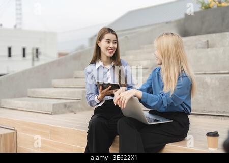 Two diverse businesswomen collaborating on a tablet and laptop while seated outdoors, sharing innovative ideas in a professional environment Stock Photo