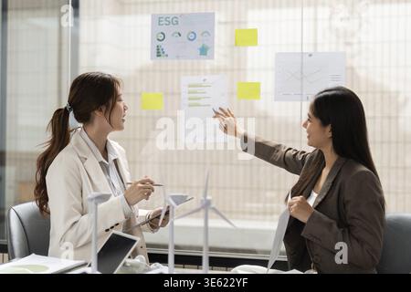 Two colleagues engage in a lively discussion about ESG insights, pointing at charts on the wall, showcasing their commitment to integrating sustainabl Stock Photo