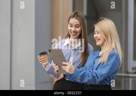 Two diverse women smiling and collaborating on a tablet while one holds a coffee, showcasing teamwork in a modern office setting Stock Photo