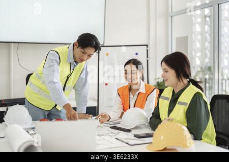 Three construction professionals, wearing safety vests and helmets, engage in a collaborative meeting to discuss and analyze building plans in a conte Stock Photo