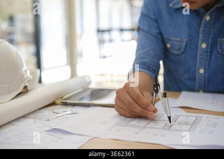Close-up of an architect's hands using a compass to draw on house plans. The desk has design tools like a tablet, pencil, and safety helmet. Bright of Stock Photo
