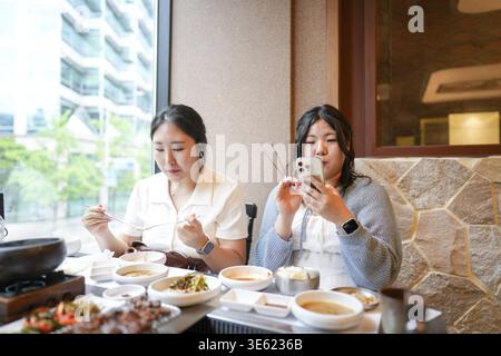 In a kalbi barbecue restaurant at a sunlit window table, two Korean women in their twenties in casual attire enjoy grilled meat and side dishes under Stock Photo