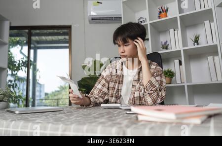 A young man sits in his home office, feeling overwhelmed as he reviews financial documents, showcasing the stress of budgeting and personal finance Stock Photo