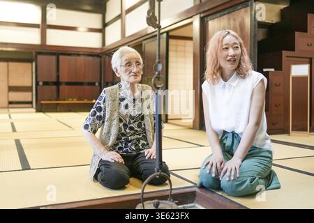Two women, one with gray hair and the other with pink hair, are sitting on a tatami mat. They are smiling at each other, creating a warm and friendly Stock Photo
