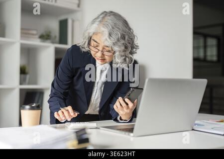 An elderly businesswoman is engaged in multitasking, using a calculator while holding a smartphone, demonstrating efficiency in her modern office spac Stock Photo