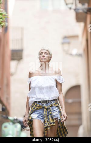 A woman wearing a white shirt and plaid shorts walks down a narrow alleyway. The scene is quiet and peaceful, with only the sound of her footsteps and Stock Photo