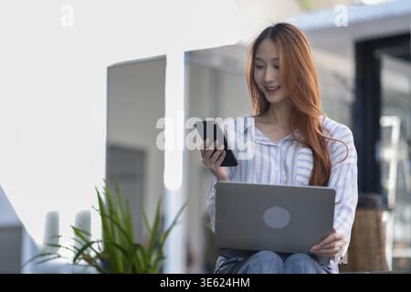 Woman is sitting on a chair and using a laptop and a cell phone. She is smiling Stock Photo
