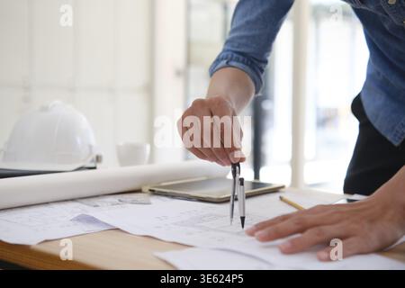 Close-up of an architect's hands using a compass to draw on house plans. The desk has design tools like a tablet, pencil, and safety helmet. Bright of Stock Photo