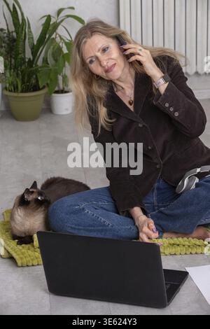Woman works from home on a laptop while talking on the phone with a cat nearby in a cozy living space Stock Photo
