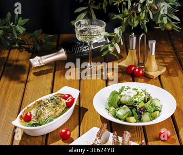 Food, A beautifully arranged table displays a gourmet meal with fresh ingredients, including a salad with greens and cucumbers, a baked dish, and a gl Stock Photo