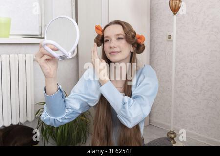 A young woman with long hair and playful curls decorates her hairstyle with bright orange clips. She gently inspects her reflection in a handheld mirr Stock Photo