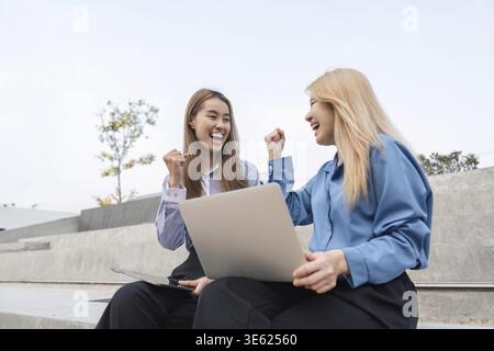 Two businesswomen outdoors joyfully celebrating their success, raising their fists in excitement while sitting with laptops on outdoor steps Stock Photo