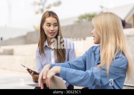 Two diverse women engaged in a friendly conversation while one holds a tablet, fostering collaboration in an outdoor environment Stock Photo