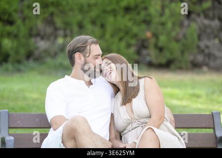 A man and a woman are sitting on a bench in a park, smiling and hugging each other Stock Photo
