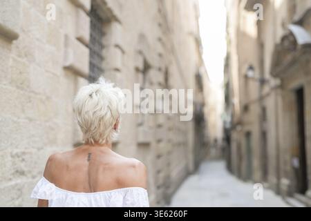 A woman in a white dress walks down a narrow alleyway. The alleyway is lined with buildings, and the woman is looking up at something. The scene has a Stock Photo