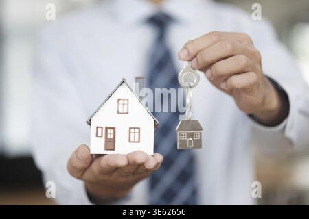 A close up of a real estate agent holding a miniature house model and a key with a house-shaped keychain, symbolizing property ownership, buying, sell Stock Photo