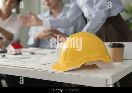 A yellow hard hat symbolizes safety in a collaborative setting, while team members discuss architectural designs and project details over blueprints a Stock Photo