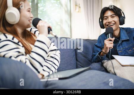 Young man recording podcast at home using microphone and headphones while speaking with co-host on sofa Stock Photo
