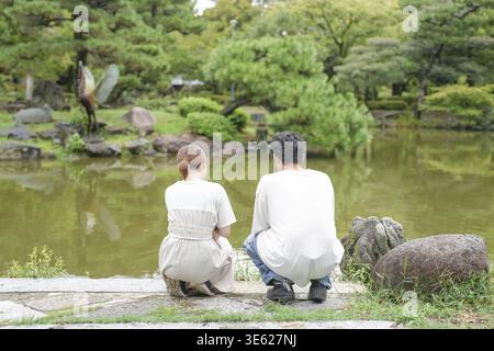 A man and woman are sitting on a stone ledge near a pond. The man is wearing a white shirt and the woman is wearing a white dress Stock Photo