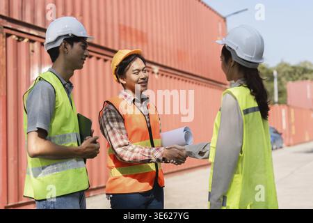 Three construction professionals engaged in a handshake, solidifying a partnership while discussing project plans near shipping containers at a constr Stock Photo