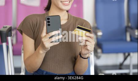 A young woman smiling while holding a smartphone and a credit card, preparing for her travel at the airport, highlighting excitement and anticipation Stock Photo