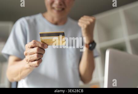 Cheerful senior man joyfully holding a credit card and making a victorious gesture after completing an online purchase in a comfortable home environme Stock Photo