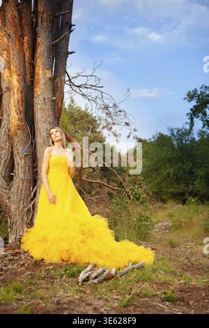 A stunning and beautiful woman in a fluffy yellow dress poses gracefully and elegantly near a large tree Stock Photo
