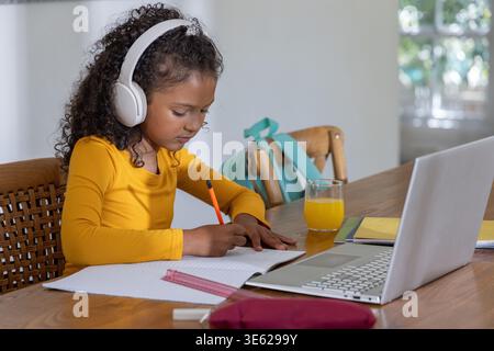 African American child wearing yellow-top headphones, studying at table, notebook and orange pencil Stock Photo