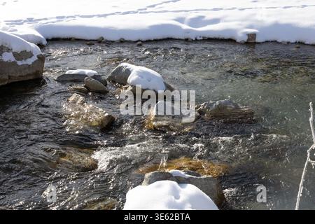 Clear winter stream flowing between snow‑covered rocks near Pontresina in the Engadin, Switzerland, showing bright alpine nature with moving water. Stock Photo