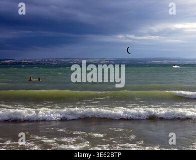 Waves rolling toward the shore under dramatic clouds with two people in the water and a kite in the distance, showing a dynamic coastal scene with moo Stock Photo