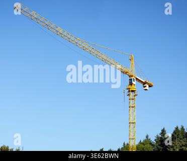 Yellow construction crane rising above green trees under a clear blue sky, showing infrastructure development in a natural forested environment. Stock Photo