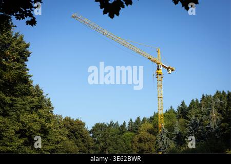 Yellow construction crane rising above green trees under a clear blue sky, showing infrastructure development in a natural forested environment. Stock Photo