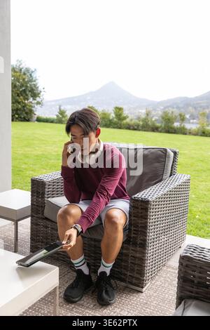 Asian man sitting on patio chair wearing maroon top holding phone pointing remote at coffee table Stock Photo