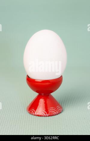 White chicken egg in red ceramic egg cup on blue green background vertical macro front view Stock Photo