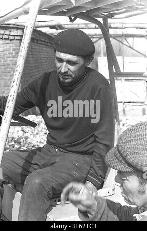 Slavyansk, 1970s, Armature-Insulator Plant. A male factory worker wearing a black beret sits in the cabin of a heavy industrial forklift parked in the outdoor manufacturing yard. He is having a brief conversation with a colleague in a flat cap standing next to the vehicle. The scene highlights the interpersonal communication and teamwork among the facility employees. An authentic historical archival photograph conveying the spirit of Soviet industrial routine everyday life, comradeship, and the atmosphere of peaceful Donbas. Stock Photo