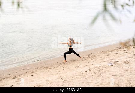 Aerial view of a fit woman practicing yoga warrior pose barefoot on a sandy river beach in autumn. Stock Photo