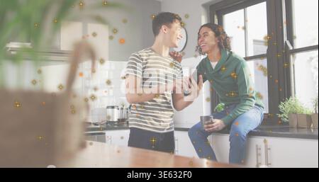 Sharing pair wearing striped tee and green hoodie, viewing smartphone in home kitchen, holding mug Stock Photo