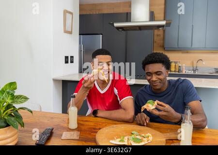 Diverse male friends sitting at kitchen table sharing pizza slices on pizza board with beverages Stock Photo