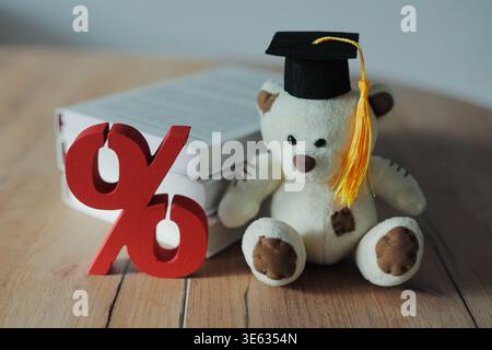 Teddy bear in graduation cap with books and percent sign on wooden table. Education, tuition fees, student loan, and academic success concept. Stock Photo