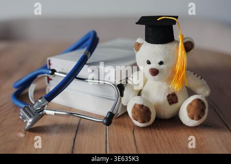 Teddy bear in graduation cap with books and stethoscope on wooden table. Medical education, healthcare training, pediatric study and student concept. Stock Photo