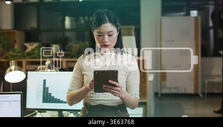 Using mid adult Asian woman in ribbed-top interacting with tablet at office, AR 37% battery overlay Stock Photo