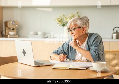 Online education courses webinar concept. Middle aged senior woman using laptop computer writing notes. Focused mature old woman enjoying studying online from home written records doing online work Stock Photo